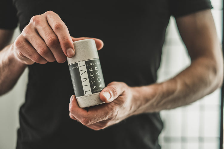 A person in a black shirt holding a cylindrical container labeled "TTVY Stick Soap" with their right hand, while their left hand rests on the lid, about to open it. The background is softly blurred and bright.