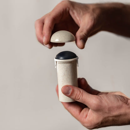 A pair of hands is holding a small beige cylindrical container with a removable lid. The person is lifting the lid to reveal a dark circular object inside, which appears to be the Grapefruit + Basil Stick Soap Refill Insert made with coconut activated charcoal. The background is plain and out of focus.