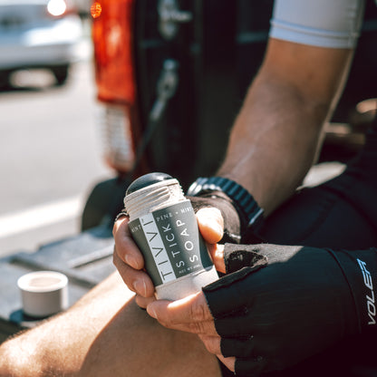 A person wearing black fingerless gloves holds a stick of Pine + Mint Stick Soap with "STIV" and "Pine, Mint" written in smaller text. Sitting outdoors with a pickup truck in the background, their other hand rests on their knee, showcasing the pure essence of essential oils in the soap while wearing a white shirt and a watch.