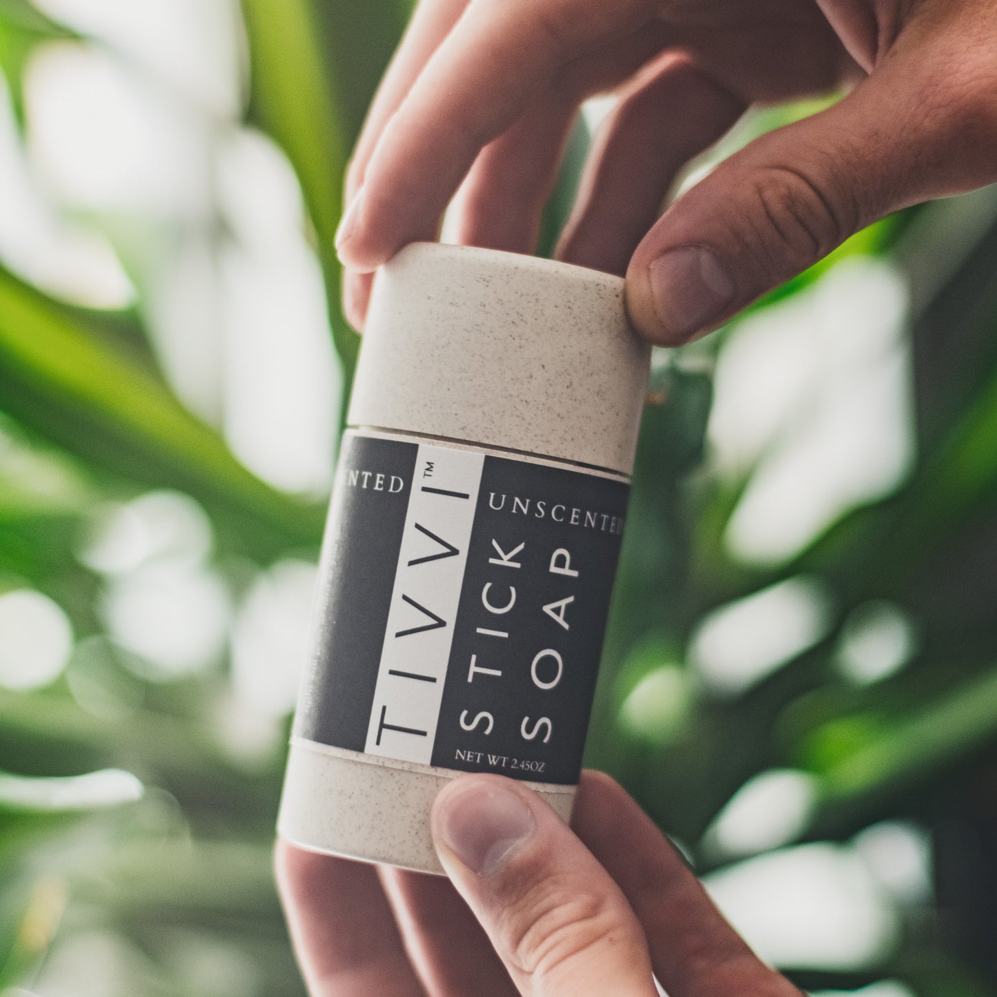 Close-up of a person holding a container of Unscented Stick Soap with both hands. The container is labeled "Unscented Stick Soap, Activated Charcoal". The background is blurred but shows green leaves, suggesting a natural or plant-filled setting.