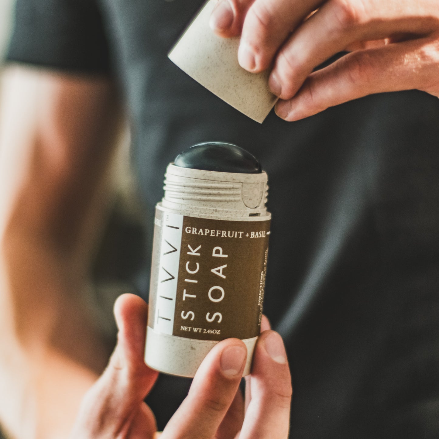 A person is holding a cylindrical container of Grapefruit + Basil Stick Soap, removing the cap with one hand. The stick soap, made with essential oils, is revealed, showing its dark tip. The label on the container includes the product name and net weight.