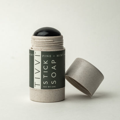 A cylindrical container in light gray, with its lid off and placed next to it, reveals a black vegan soap stick inside. The label on the container reads "TIVVI Sticks Soap" in the flavor "Pine + Mint.