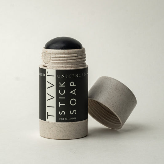 A cylindrical stick of vegan soap, featuring a black rounded top and housed in a speckled grey container, with the label reading "Unscented Stick Soap." The container lid is placed on its side next to the soap on a plain white background. The formula includes coconut activated charcoal for a deep cleanse.