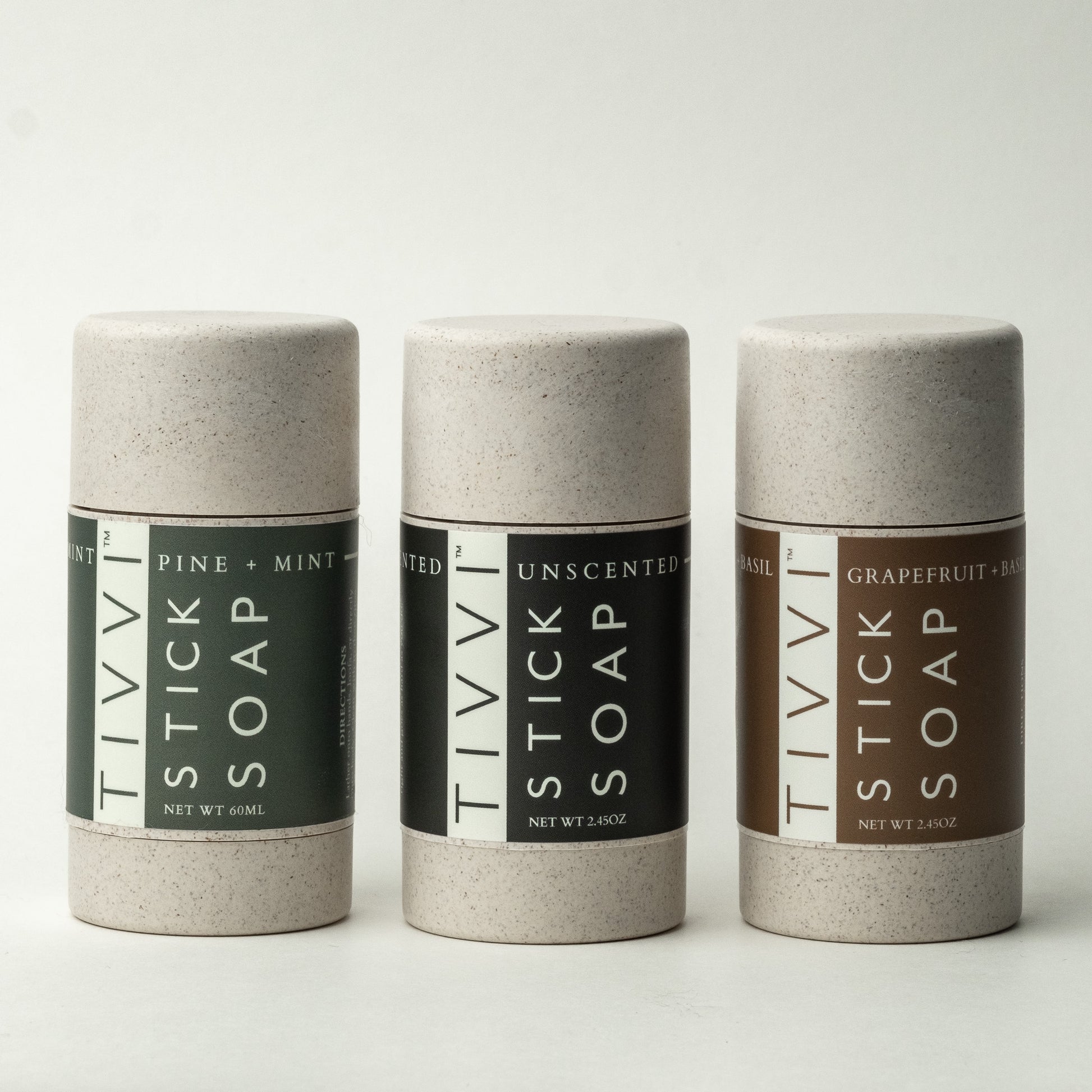 Three cylindrical soap containers are displayed against a white background. Each container is labeled "Stick Soap Tri-Pack" with different scents: Pine + Mint, Unscented, and Grapefruit + Basil. The labels are color-coded green, white, and brown, respectively. Try our Vegan Stick Soap Tri-Pack for a cleaner experience.