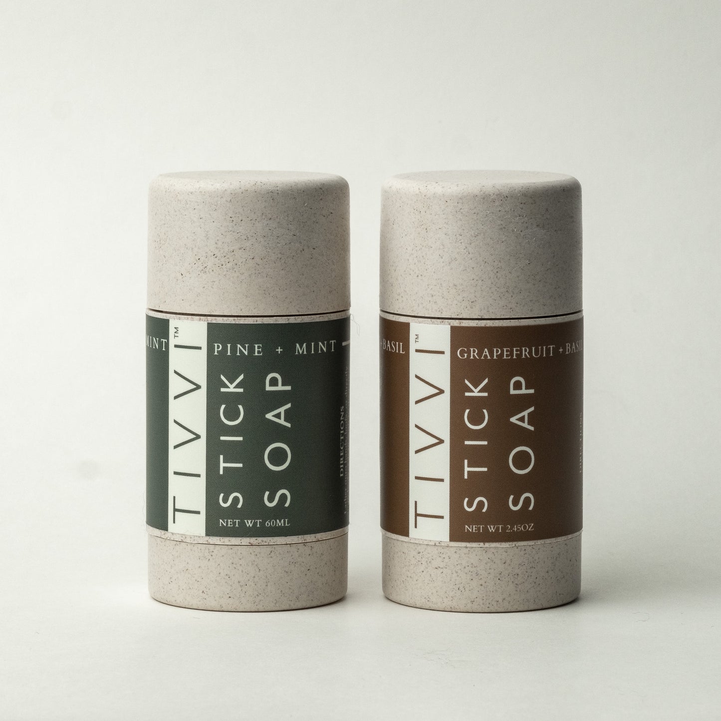 Two cylindrical packages from the "Stick Soap Scented Duo-Pack" are displayed. One soap is a green-gray color labeled "TIVVI STICK SOAP PINE + MINT," and the other is a brown color labeled "TIVVI STICK SOAP GRAPEFRUIT + BASIL." Both vegan soaps exude a minimalistic design, with the green-gray containing 60mL and the brown containing 2.84oz.