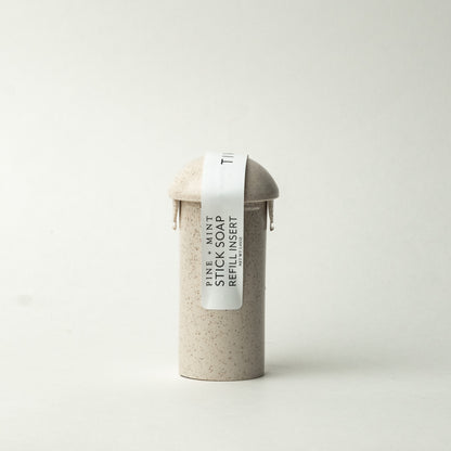 A cylindrical, beige container with a domed lid stands against a plain white background. A white label wraps around the upper part, displaying the text "Pine + Mint Stick Soap Refill Insert". The surface of the container features small speckles. This product is a vegan soap with sustainable refill inserts, named Pine + Mint Stick Soap Refill Insert.