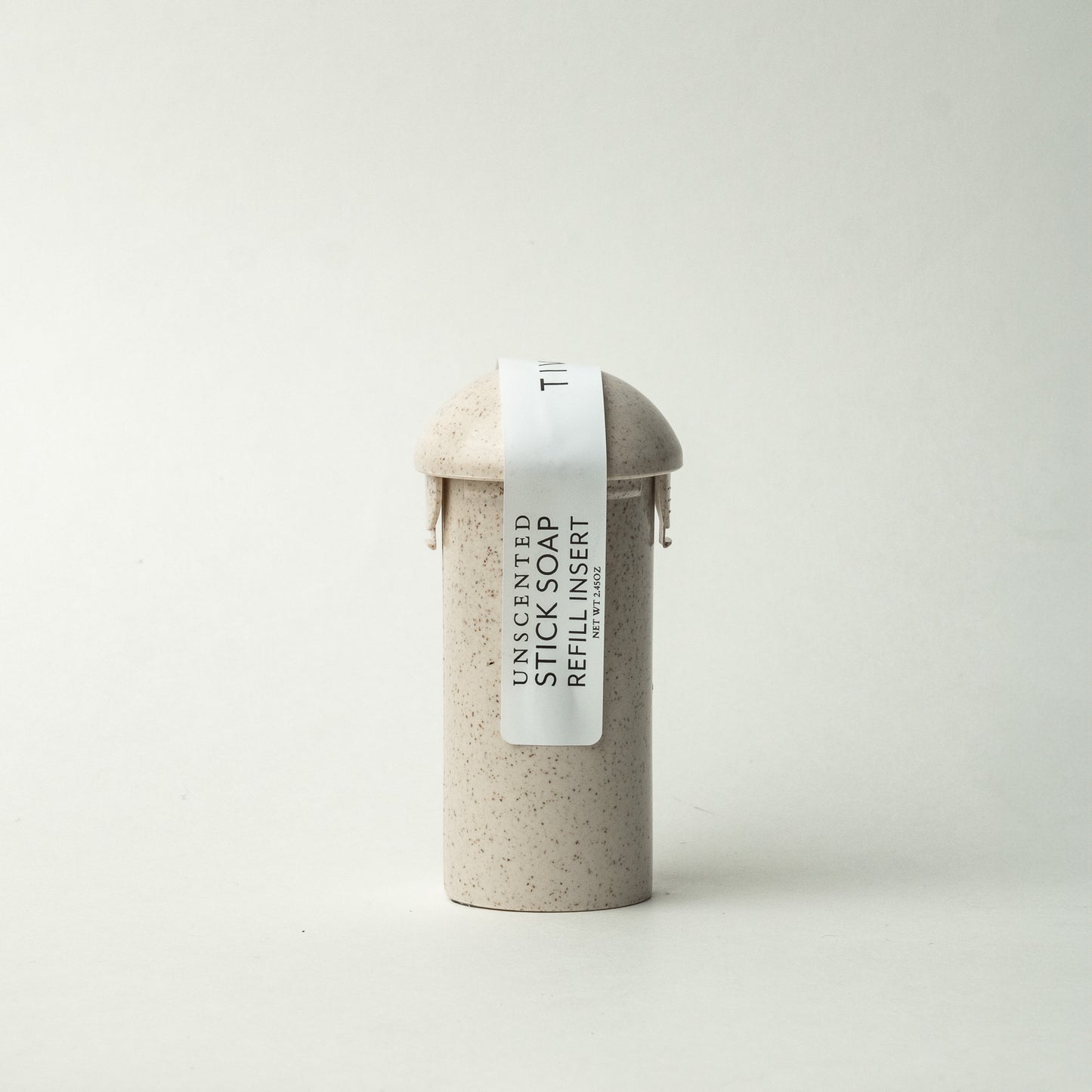 A minimalist cylindrical container with a rounded top is displayed against a plain background, labeled "Unscented Stick Soap Refill Insert" on a vertical white paper strip. The vegan soap, featuring a simple and speckled off-white design, contains coconut activated charcoal.