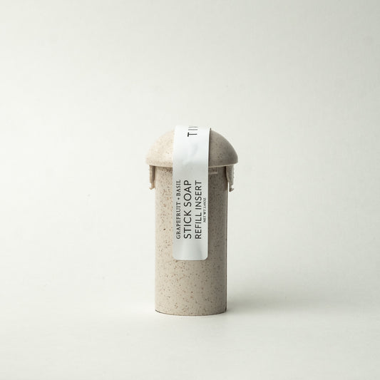 A beige cylindrical container with a domed lid, labeled "Grapefruit + Basil Stick Soap Refill Insert" in black text on a white ribbon-like strip that extends vertically down the front. The vegan soap features coconut activated charcoal for a gentle cleanse. The background is plain white.