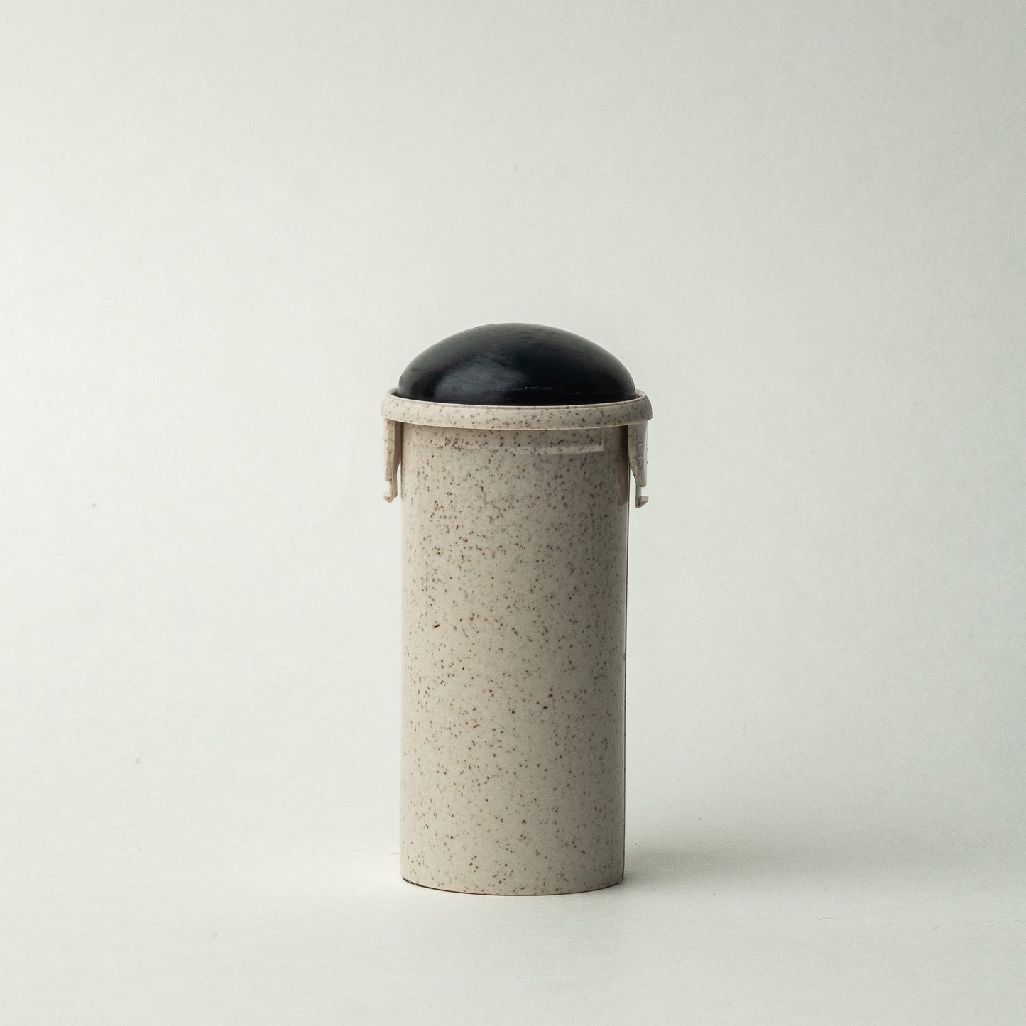 The Pine + Mint Stick Soap Refill Insert is elegantly designed as a small, cylindrical container with a speckled beige body and a rounded black lid. Its simple, minimalist design makes it ideal for holding coconut activated charcoal or vegan soap, offering a sustainable option. The black lid fits snugly on top against the plain, light gray background.